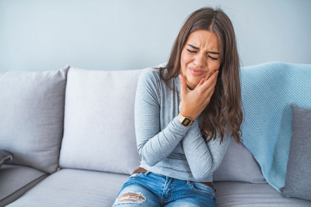 Woman on grey couch holding hands to jaw in pain